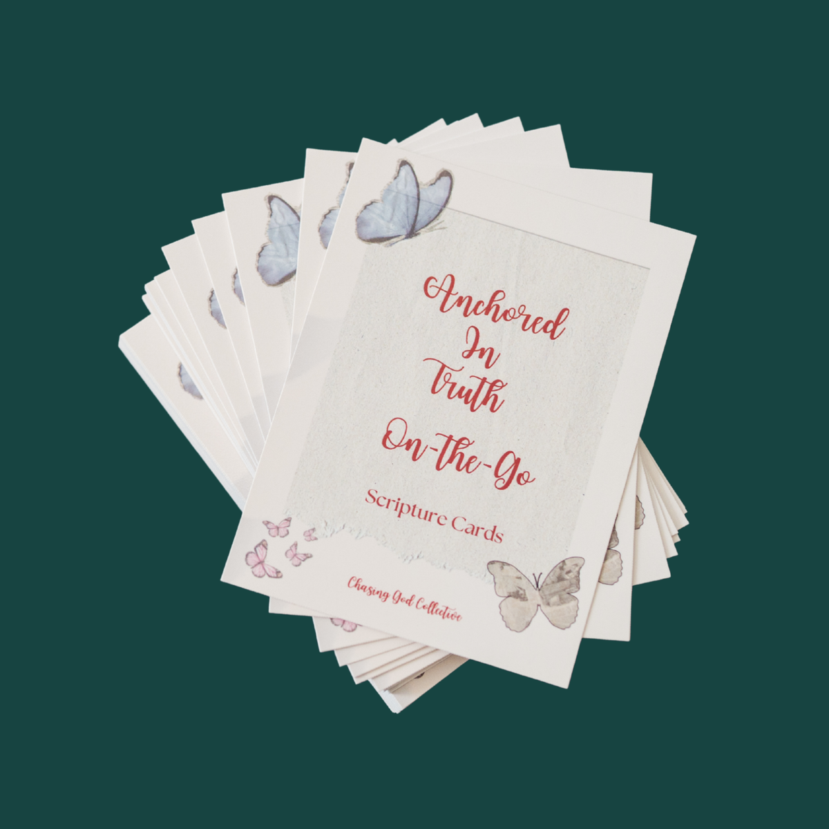A fan of scripture cards from the 'Anchored In Truth On-the-Go' collection by Chasing God Collection, displayed on a solid dark teal background. The cards are cream-colored with watercolor butterfly illustrations and red script text.