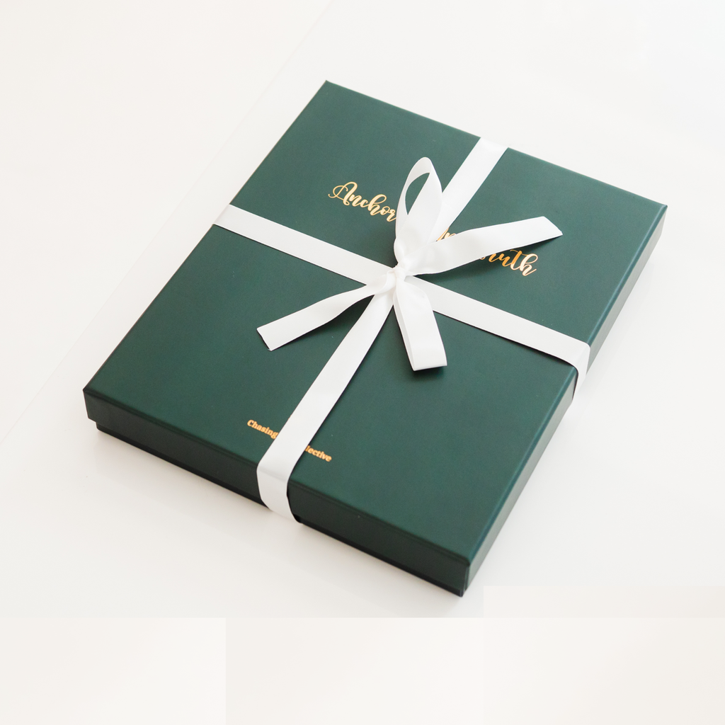 A dark green rectangular gift box tied with a white ribbon and bow, featuring gold foil script text on the lid, photographed on a clean white background.