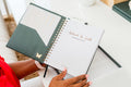 An open spiral-bound journal or planner with a dark green cover and white inner pages, held by a person in a bright indoor setting. The right page has gold foil text reading 'Adopted In Truth' and a small butterfly graphic on the cover.