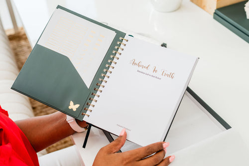 An open spiral-bound journal or planner with a dark green cover and white inner pages, held by a person in a bright indoor setting. The right page has gold foil text reading 'Adopted In Truth' and a small butterfly graphic on the cover.