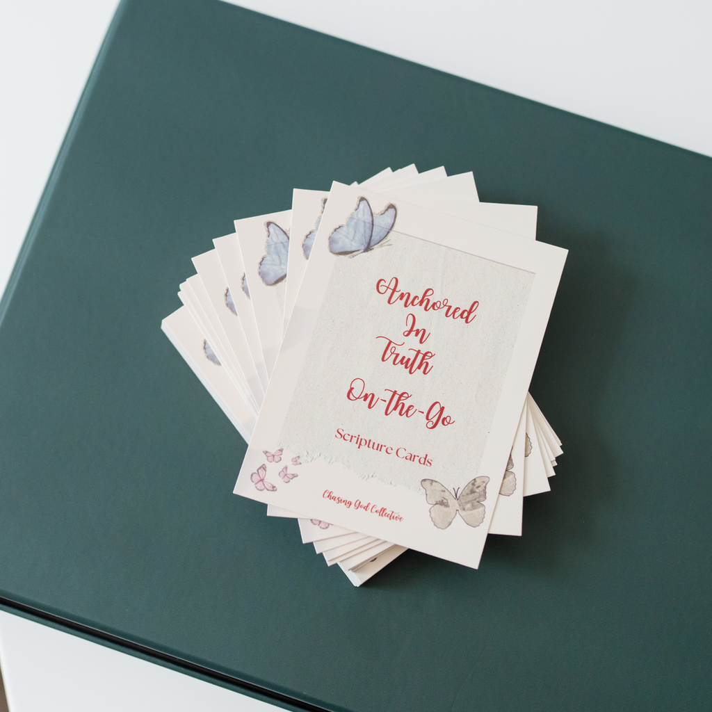 A stack of scripture cards fanned out on a dark teal notebook. The cards are off-white with red script text reading 'Anchored In Truth On-The-Go Scripture Cards' and decorated with watercolor butterflies in blue, pink, and beige.