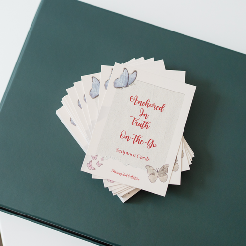 A stack of scripture cards fanned out on a dark teal notebook. The cards are off-white with red script text reading 'Anchored In Truth On-The-Go Scripture Cards' and decorated with watercolor butterflies in blue, pink, and beige.