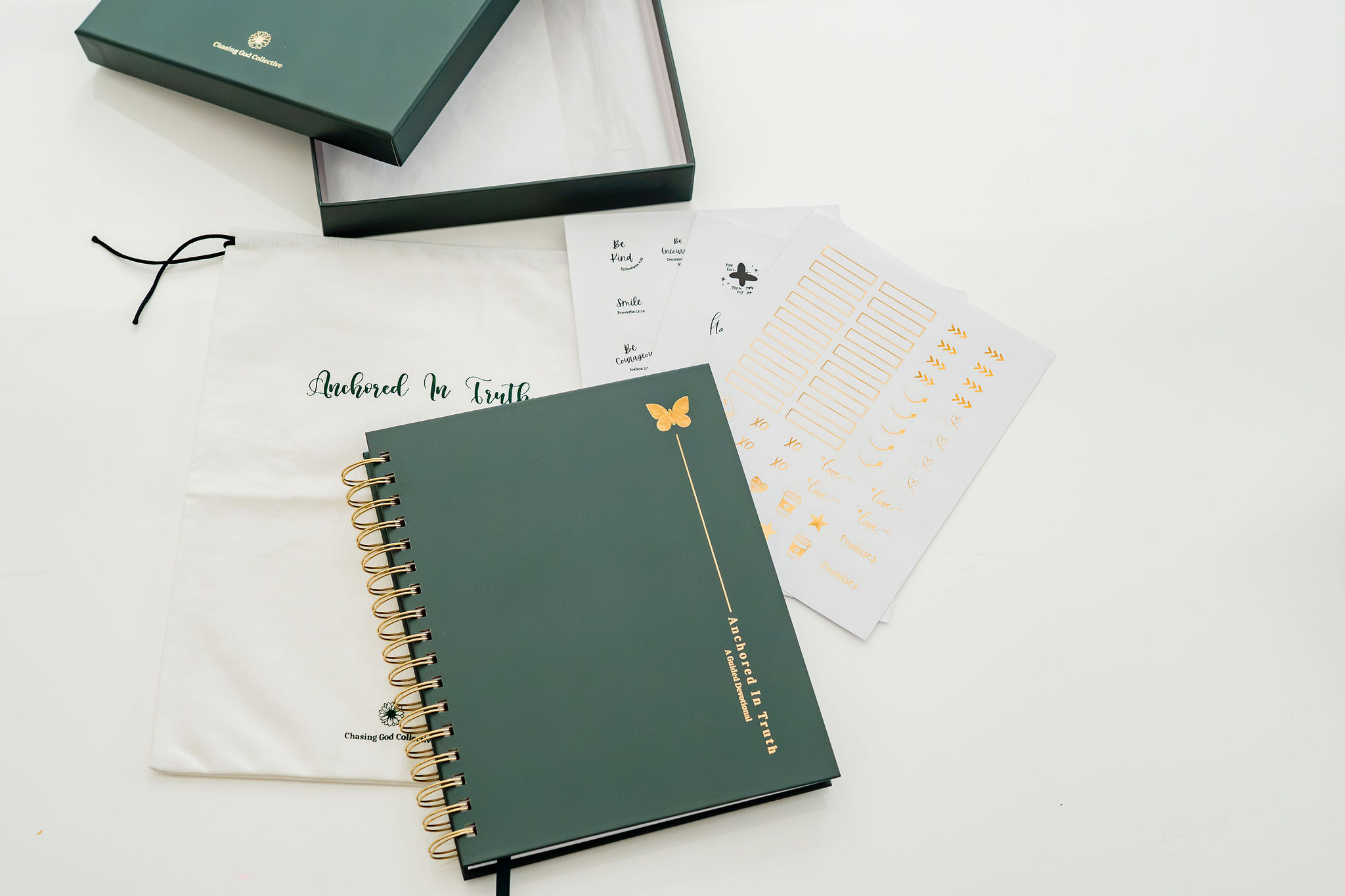A dark green spiral-bound devotional with gold hardware and a butterfly emblem, displayed with its white dust bag, open presentation box, and printed stickers on a clean white background.