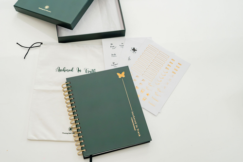 A dark green spiral-bound devotional with gold hardware and a butterfly emblem, displayed with its white dust bag, open presentation box, and printed stickers on a clean white background.